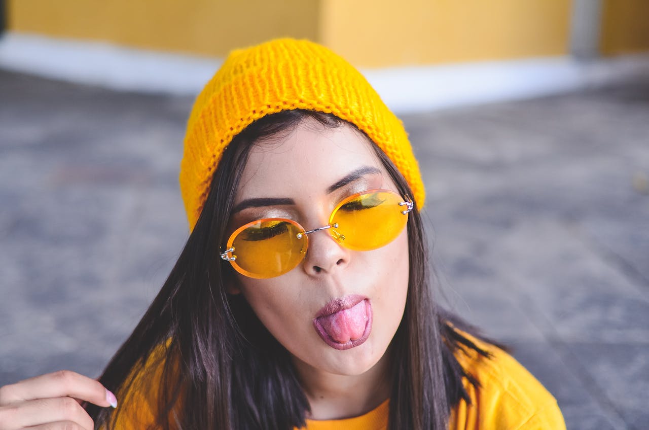 about-01 A young woman in stylish yellow attire sticks out her tongue in a playful gesture outdoors.